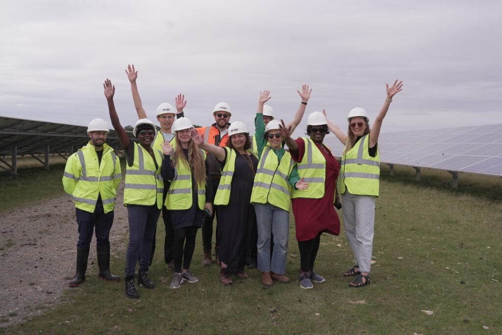 Members of the community steering group and BD Giving staff are all wearing high-vis jackets and hard hats. They're standing in an empty field on a cloudy day, waving at the camera. Everyone looks happy.