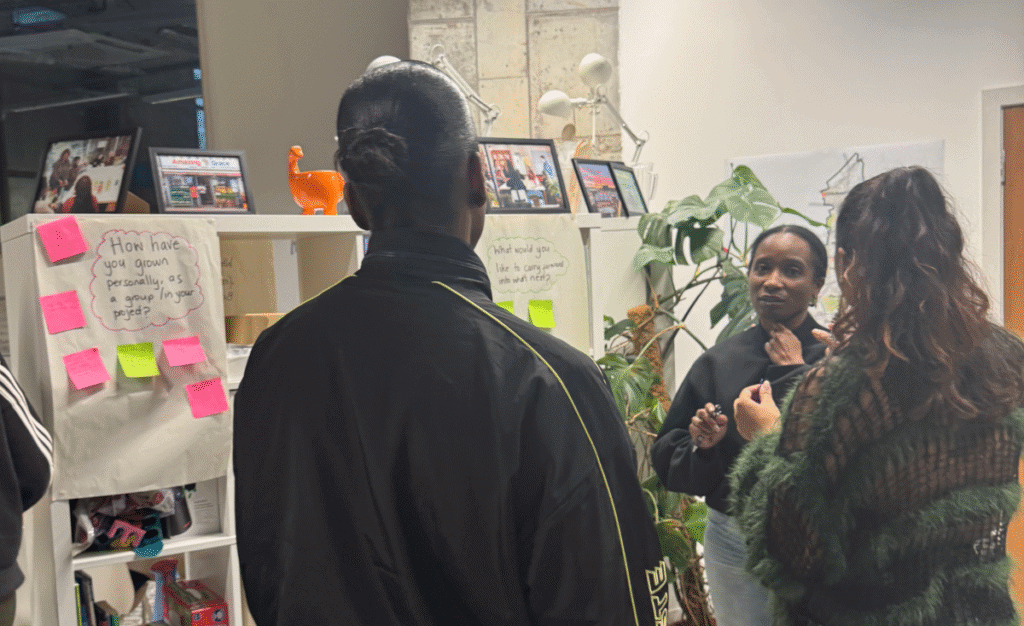 3 women are talking while standing in an office environment. 2 of them have their back to the camera.