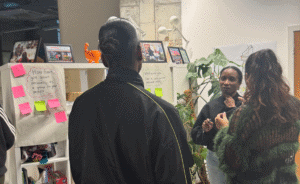 3 women are talking while standing in an office environment. 2 of them have their back to the camera.
